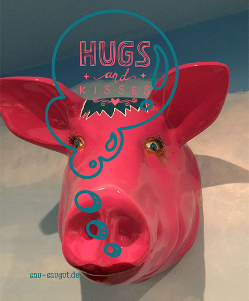 Ein an der Wand hängender Sauenkopf in der Farbe pink. Schriftzug als Sprechblase: Hugs and Kisses und unterhalbt sau-saugut.de 