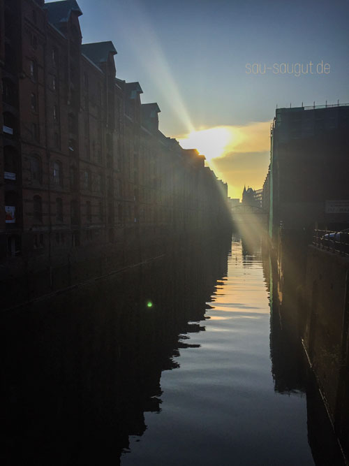 Sonnenaufgang im historischen Hafen in Hamburg. Man sieht die Lagerhäuser entlang der Wasserstraße. Schriftzug oberhalb sau-saugut.de